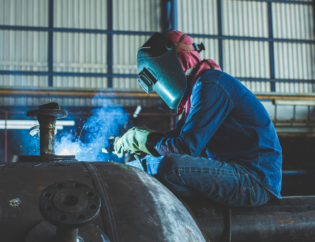 A welder working on a large steel structure inside a fabrication workshop, demonstrating industrial steelwork and connection techniques used in modern construction